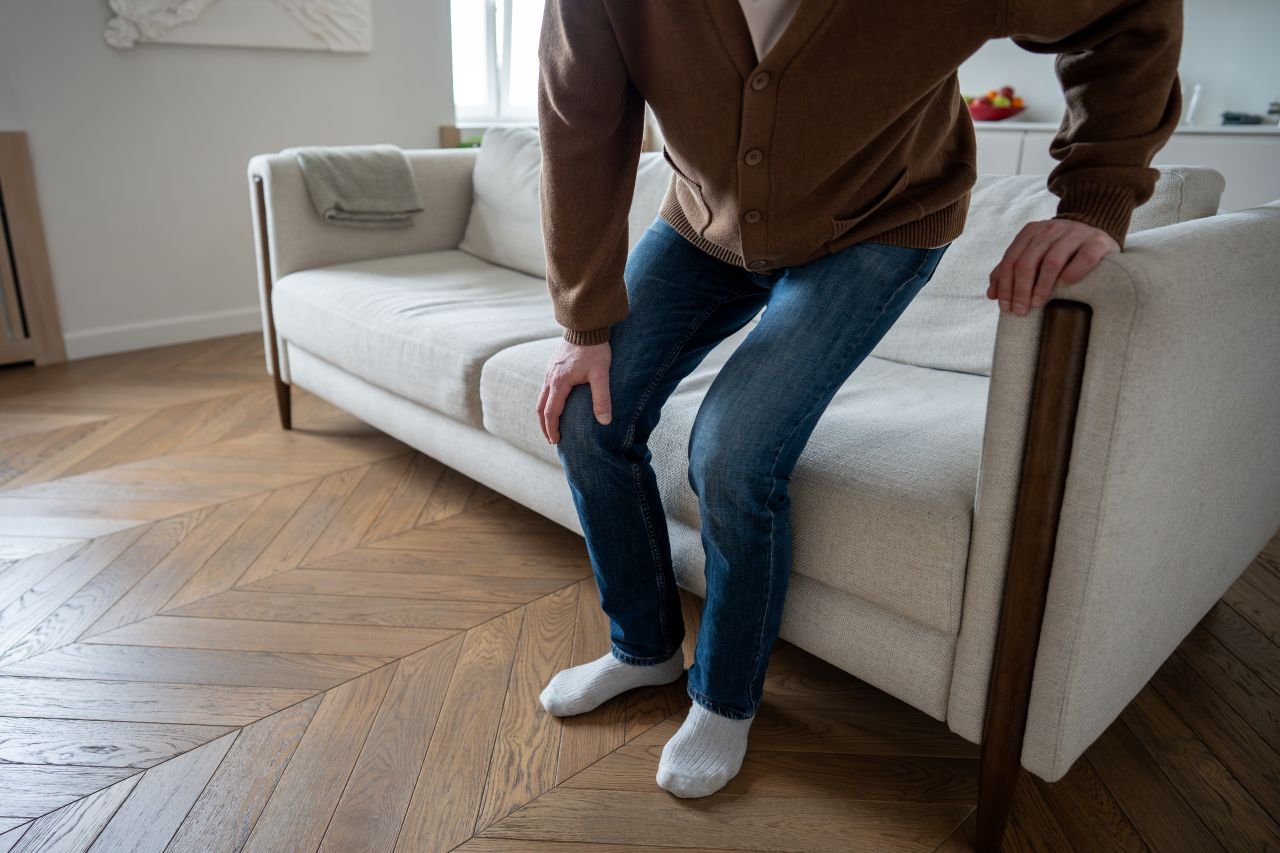 Adult man over 55 sitting in a low deep sofa showing how low seat height increases knee demand when getting up from a chair with knee pain.