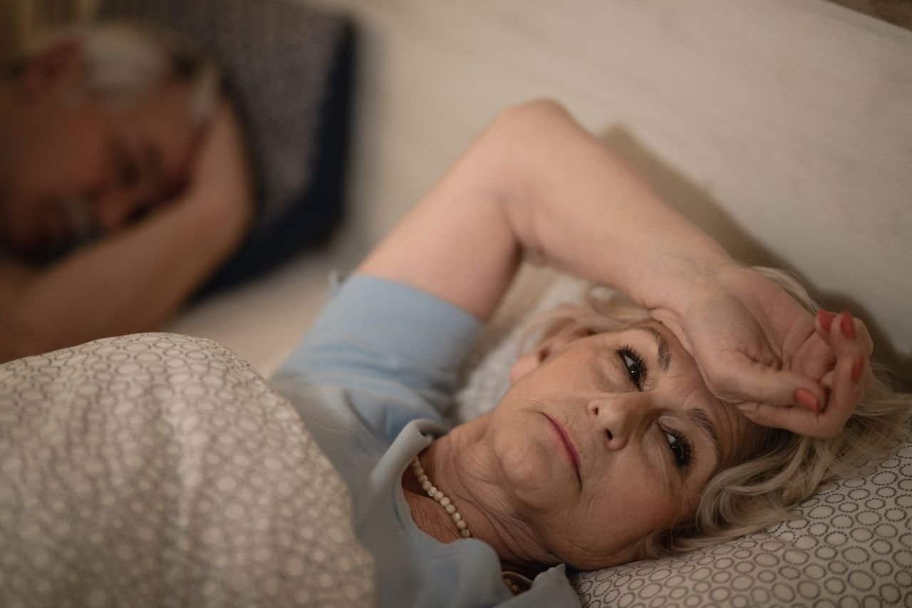 Adult woman over 55 lying awake in bed in the early morning showing the sleep disruption that feeds the inflammation loop affecting daily walking capacity.