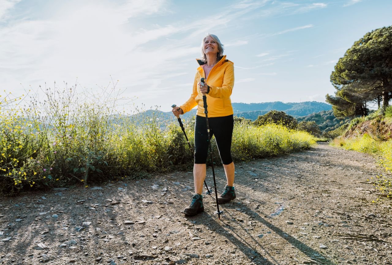 Adult woman over 60 using walking poles on an uneven path showing how poles address the balance deficit that knee OA creates during walking.