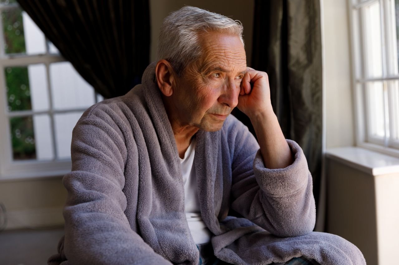Adult man over 55 sitting indoors on a rainy day in an armchair illustrating the inactivity spiral that compounds knee pain when movement stops.