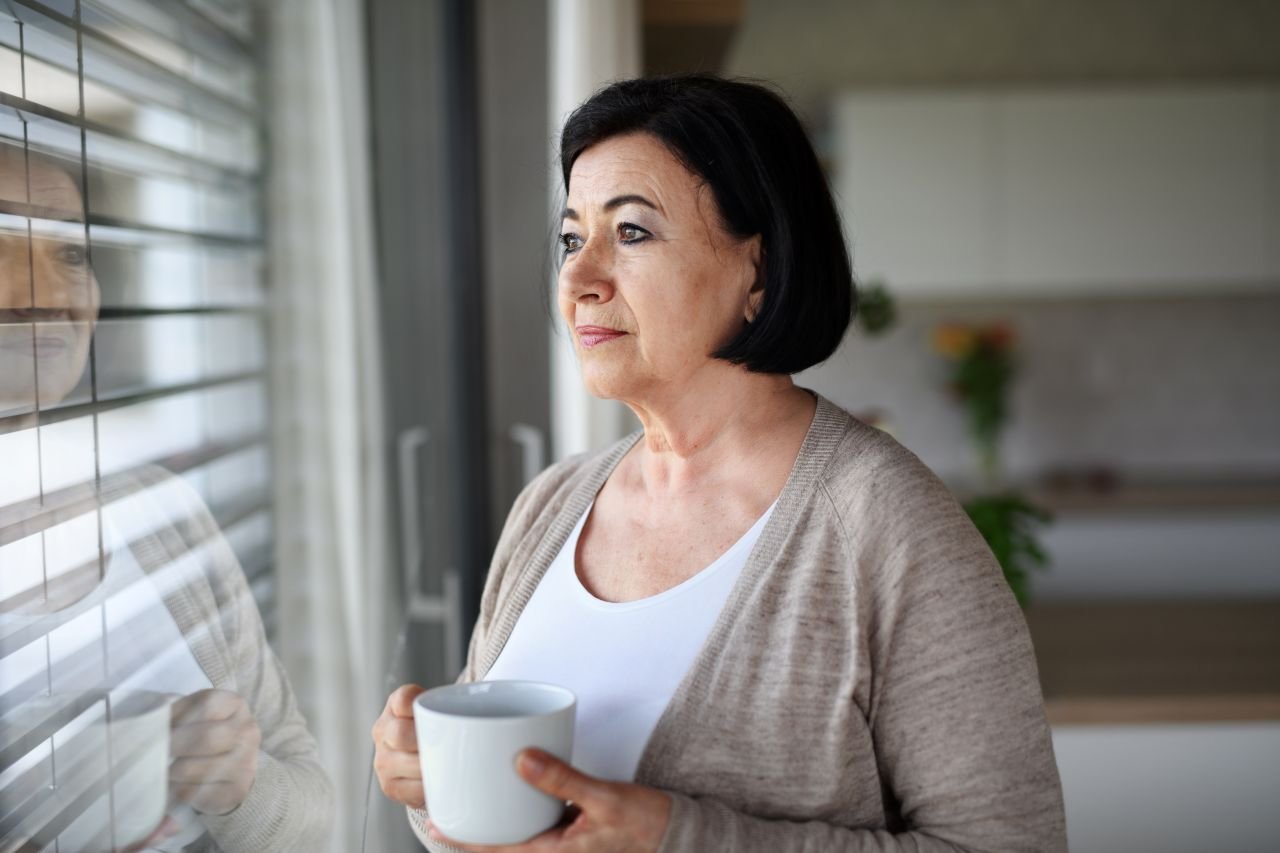 Adult woman over 55 holding a warm mug on a rainy morning reflecting on why some adults feel weather-related knee pain more than others.